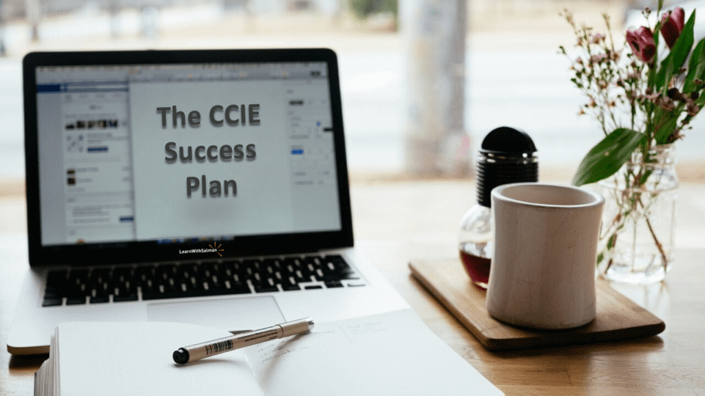 The_CCIE_Success_Strategy_Plan A laptop displaying the text “The CCIE Success Plan” sits on a wooden table next to an open notebook, a pen, a white coffee mug on a saucer, and a small vase with flowers. The scene is set near a window with a blurred view outside.